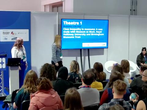 A photograph showing two women presenting an event to around thirty people.